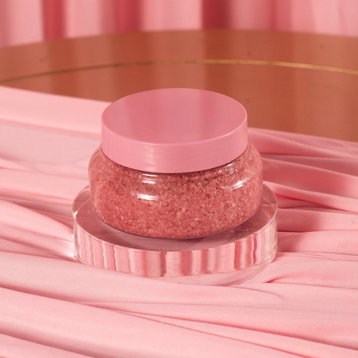 A clear jar of Viva La Bombshell Burlesque's Diamond Salt Soak, with a pink lid and filled with pink Dead Sea salt scrub, sits on a round acrylic stand, surrounded by soft pink fabric and a matching pink background.