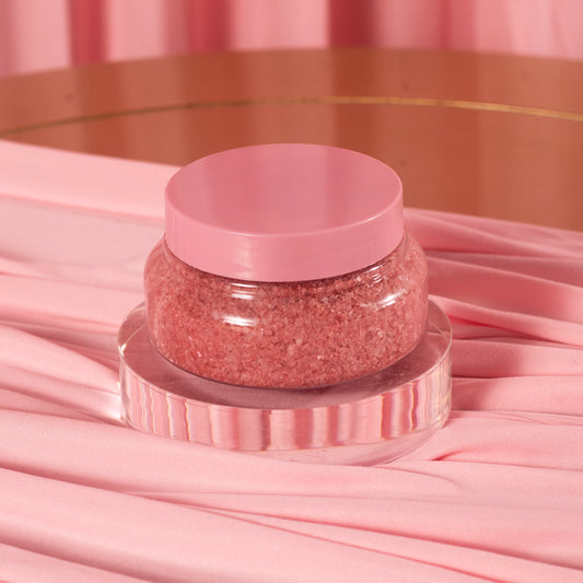 A clear jar of Viva La Bombshell Burlesque's Diamond Salt Soak, with a pink lid and filled with pink Dead Sea salt scrub, sits on a round acrylic stand, surrounded by soft pink fabric and a matching pink background.