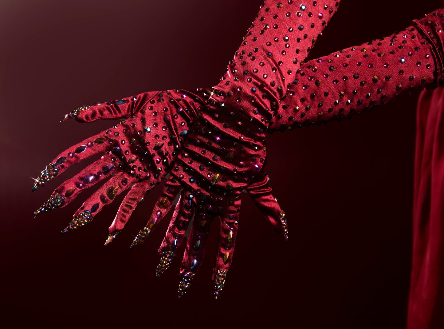 Close up of burgundy nail gloves with multi-color rhinestone accents on a dark background. Both hands with fingers spread to show details. 