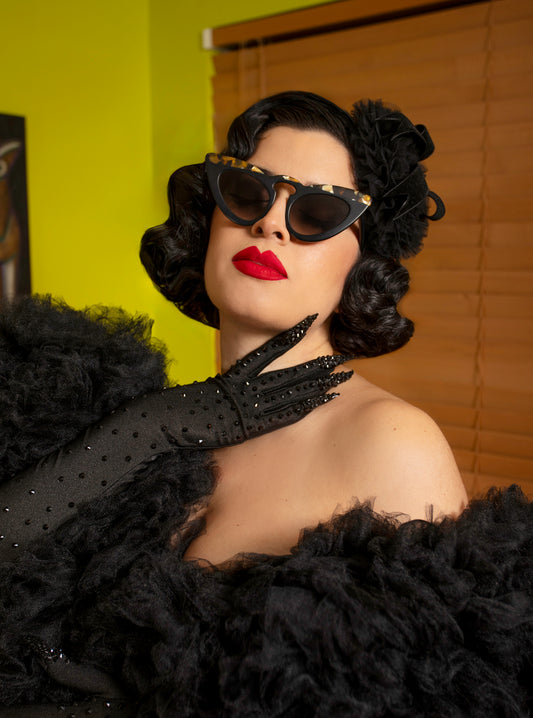 Female with black hair, sunglasses, and red lipstick against a yellow and brown wall wearing black rhinestone .ail gloves
