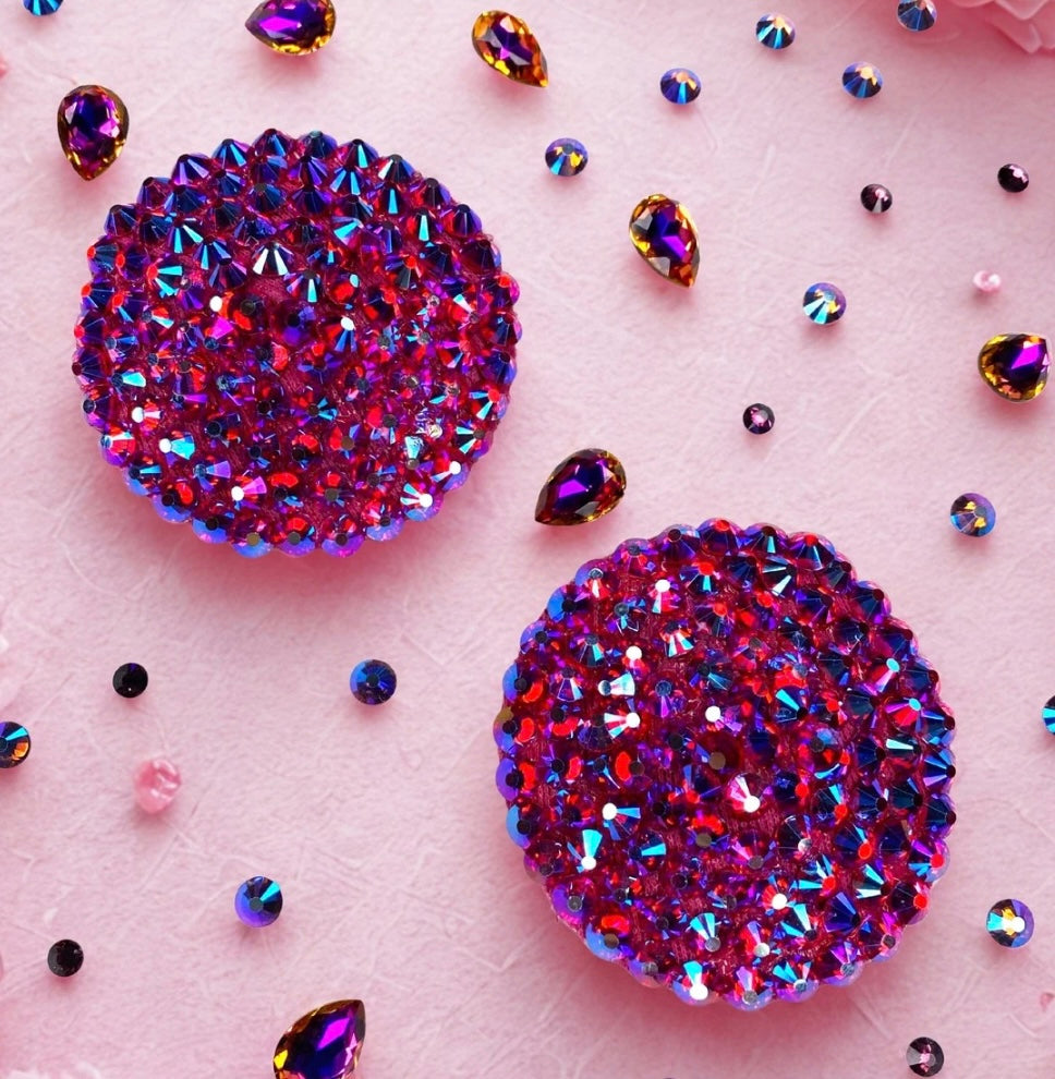 Two Simple Rhinestone Pasties (Circle) by Viva La Bombshell Burlesque, featuring sparkling purple and pink gemstones, rest on a light pink surface with scattered colorful gem-like decorations.