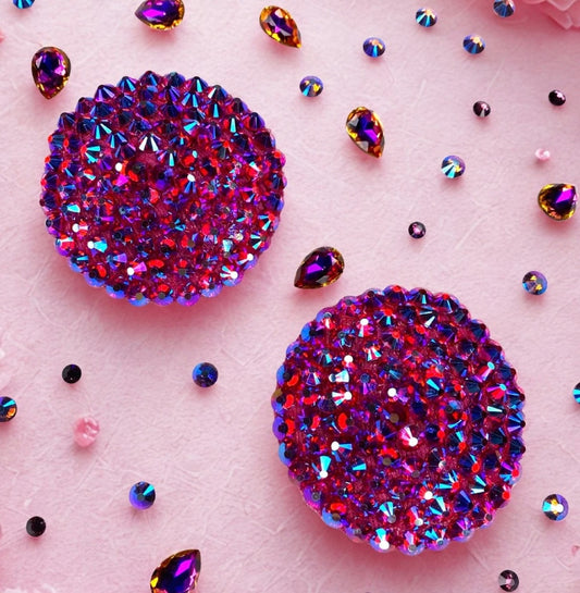 Two Simple Rhinestone Pasties (Circle) by Viva La Bombshell Burlesque, featuring sparkling purple and pink gemstones, rest on a light pink surface with scattered colorful gem-like decorations.
