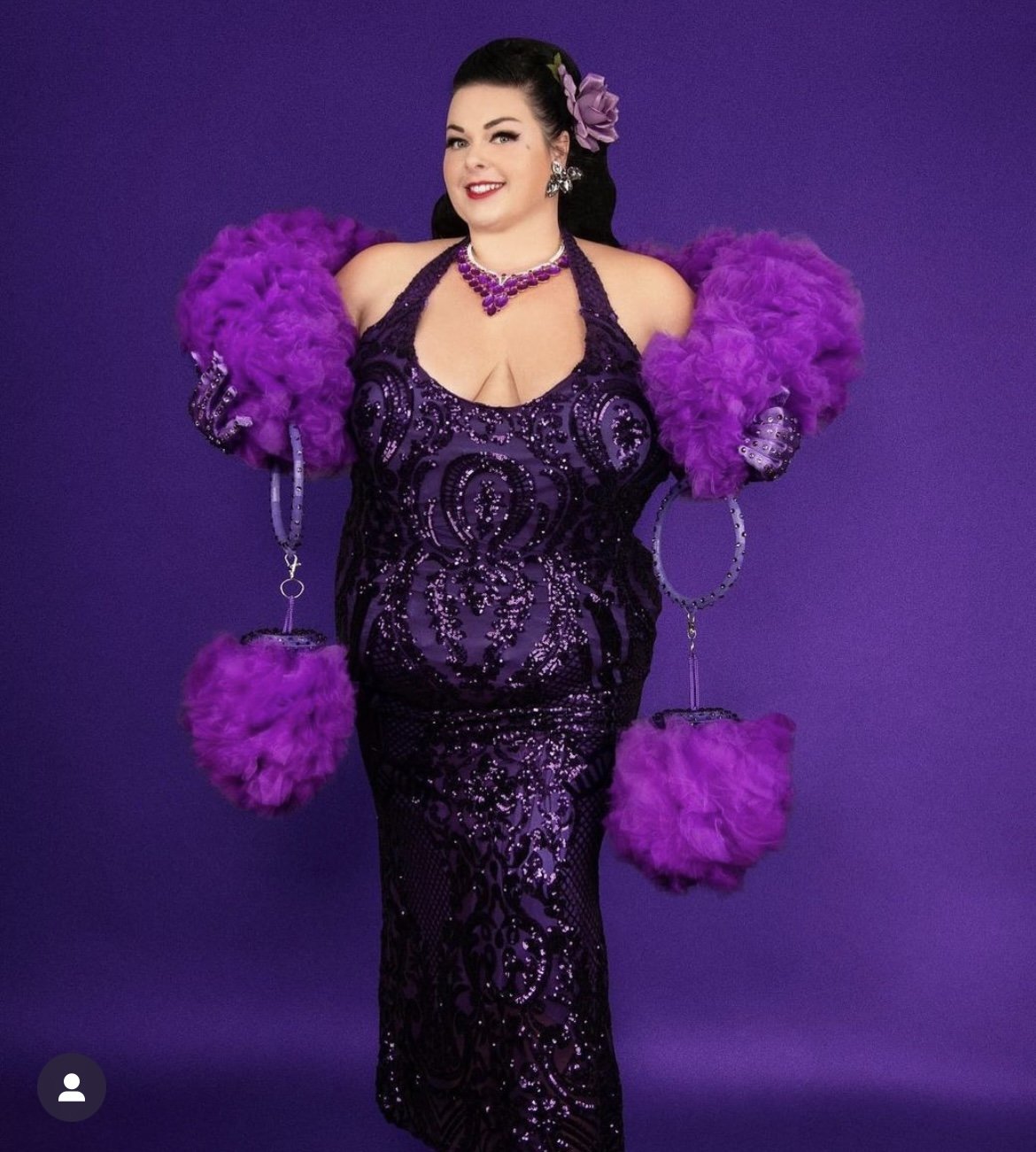 A woman poses against a purple background in a sparkling dark purple gown, fluffy gloves, and The De'Vil - Tulle Boa by Viva La Bombshell Burlesque. She holds feathered props and wears a custom necklace, earrings, and a floral hair accessory.