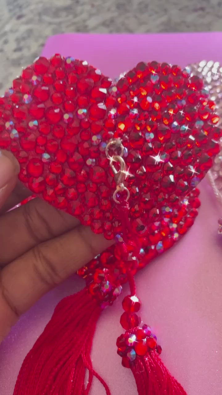 A hand holding a red  rhinestoned pasties with red tassels. Pink rhinestoned pasties with pink tassels, shown next to it on a countertop.