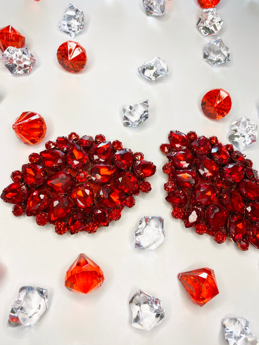 Juicy Pasties by Viva La Bombshell Burlesque, featuring LT Siam red gemstones and clear stones, are displayed on a white surface. Two large clusters of red gems sit center stage, surrounded by scattered individual stones.