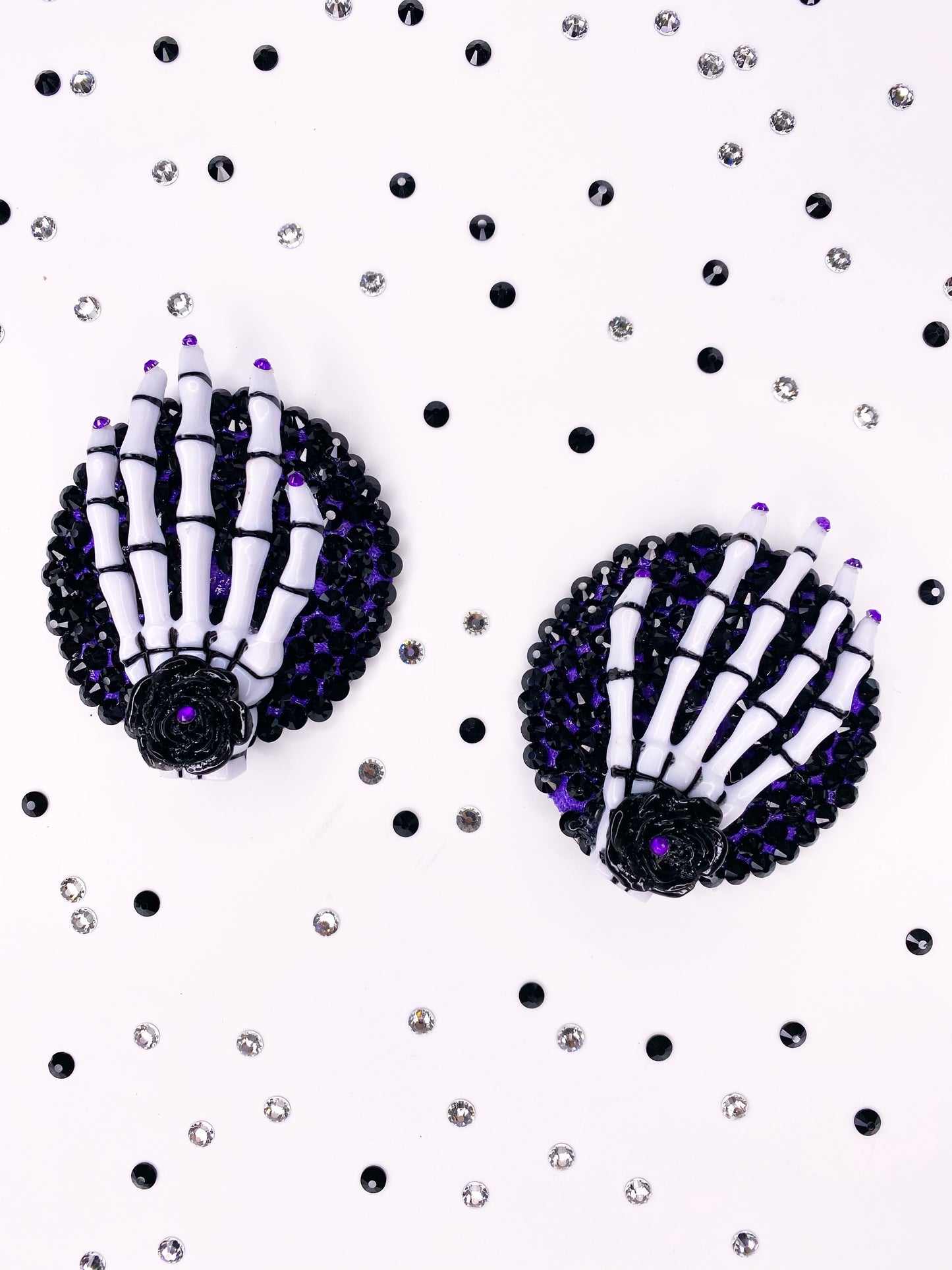 A pair of white and black skeleton hand pasties with rhinestone purple nails and a black flower with purple rhinestone in the center of the flower. The base is painted purple underneath black rhinestones. 