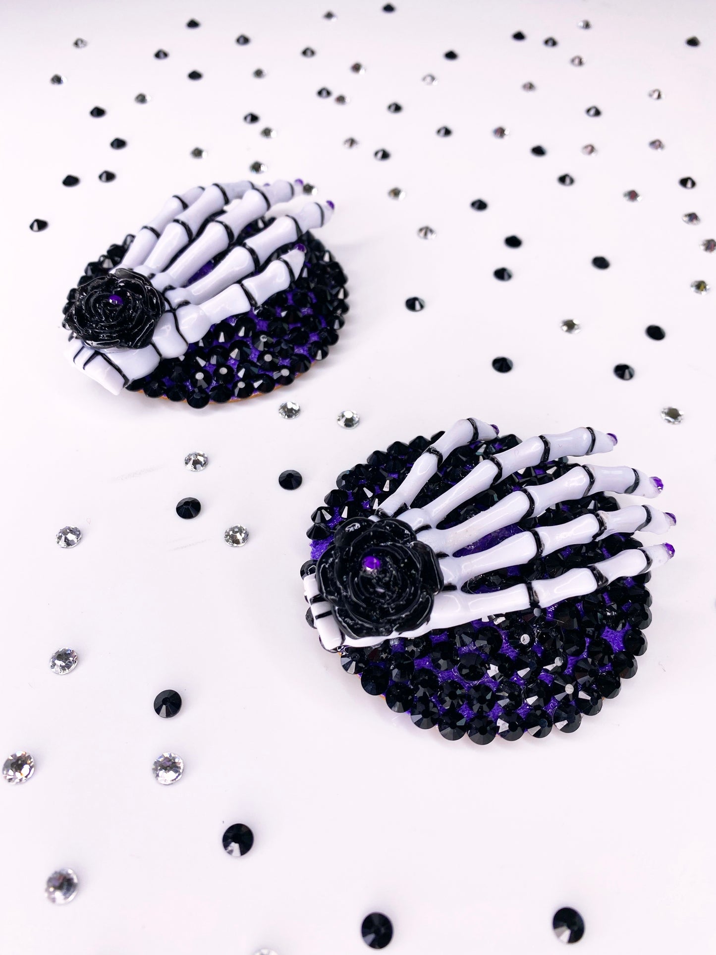 A closer up photo of a pair of white and black skeleton hand pasties with rhinestone purple nails and a black flower with purple rhinestone in the center of the flower. The base is painted purple underneath black rhinestones. 