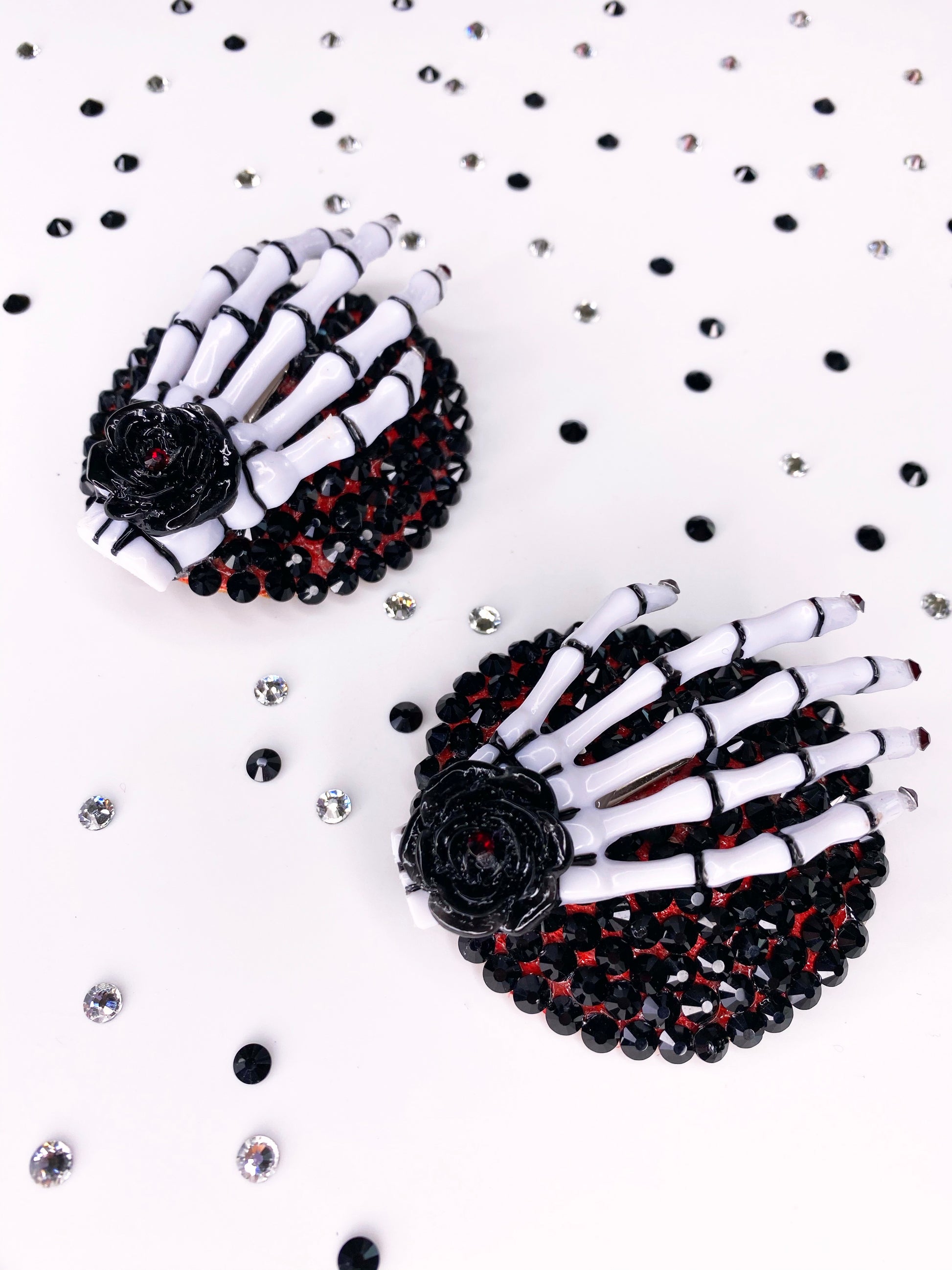 A pair of white and black skeleton hand pasties with rhinestone red nails and a black flower with red rhinestone in the center of the flower. The base is painted red underneath black rhinestones. 