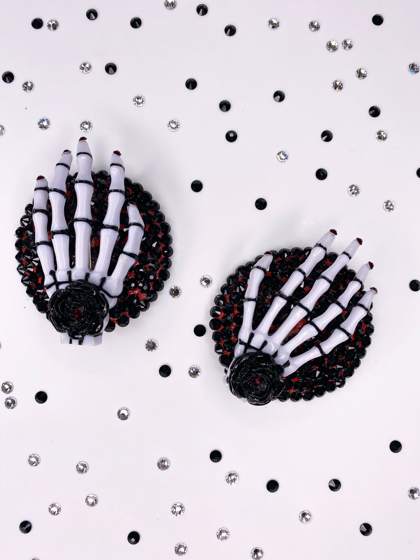A close photo of a pair of white and black skeleton hand pasties with rhinestone purple nails and a black flower with purple rhinestone in the center of the flower. The base is painted purple underneath black rhinestones. 