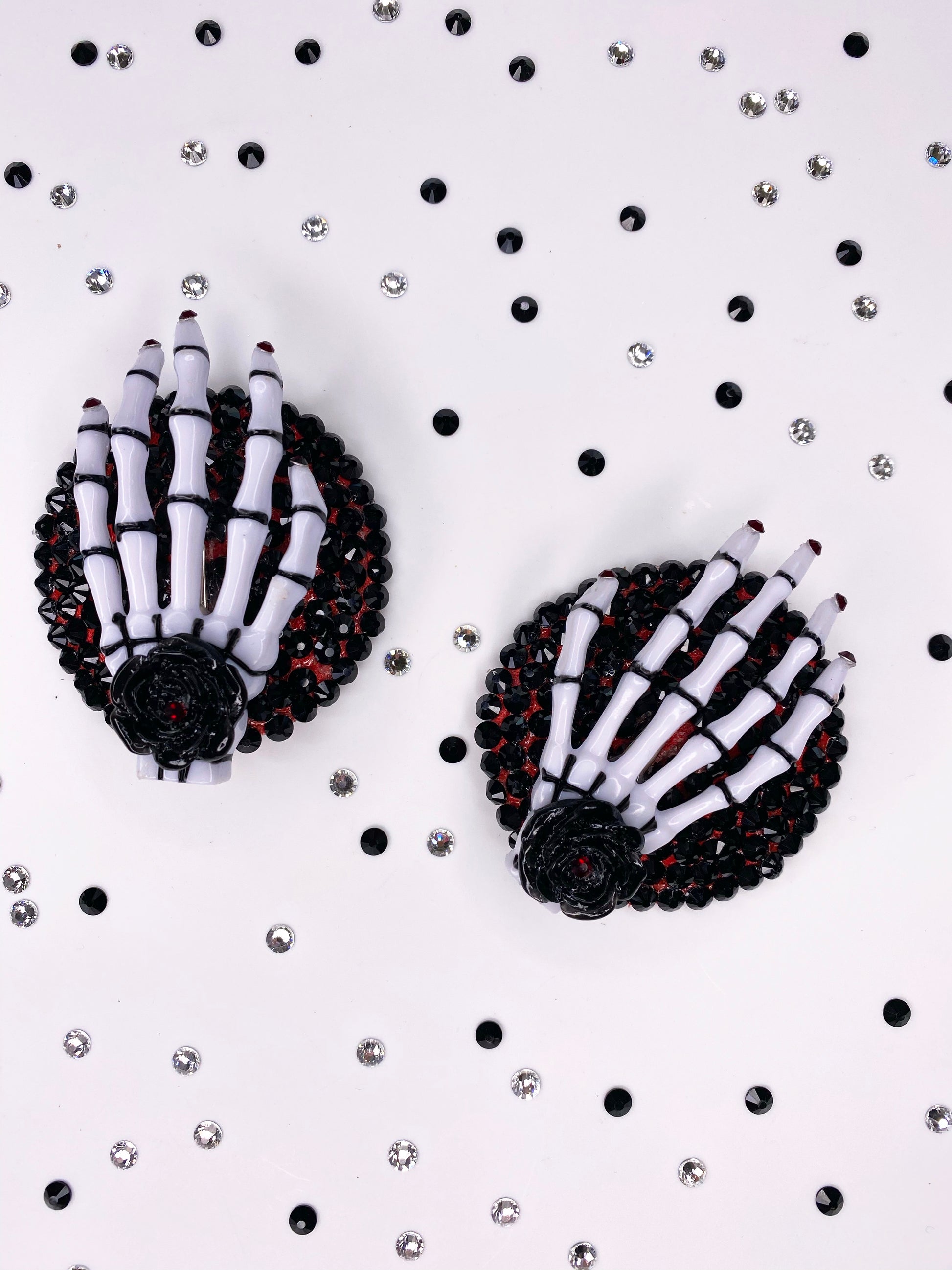 A close photo of a pair of white and black skeleton hand pasties with rhinestone purple nails and a black flower with purple rhinestone in the center of the flower. The base is painted purple underneath black rhinestones. 