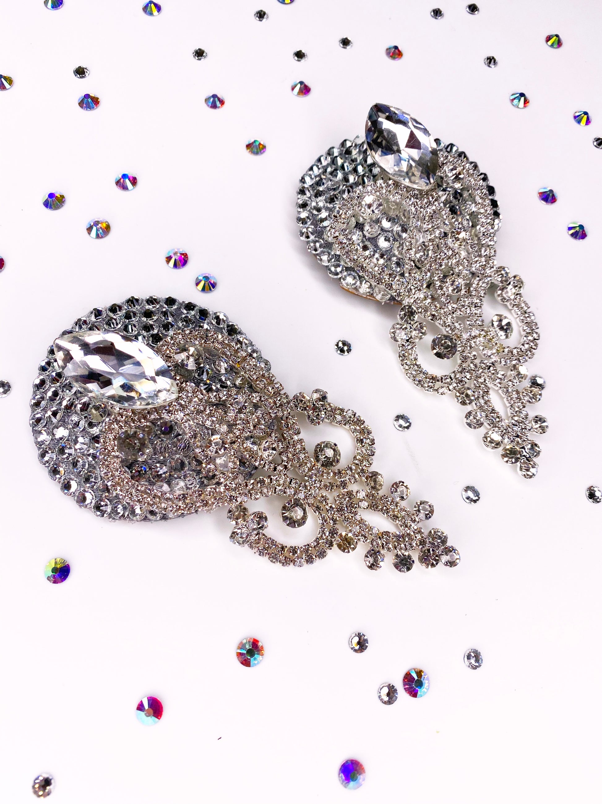 Two ornate Showgirl Pasties by Viva La Bombshell Burlesque, featuring large teardrop gems, rest on a white surface scattered with iridescent crystal rhinestones.