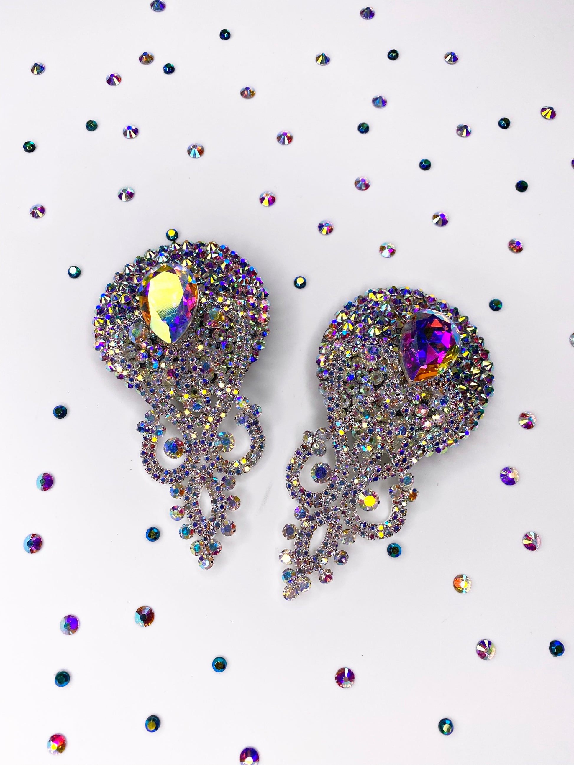 Two ornate Showgirl Pasties by Viva La Bombshell Burlesque, featuring iridescent jewels and intricate designs, are displayed on a white surface surrounded by scattered crystal rhinestones of varying sizes.