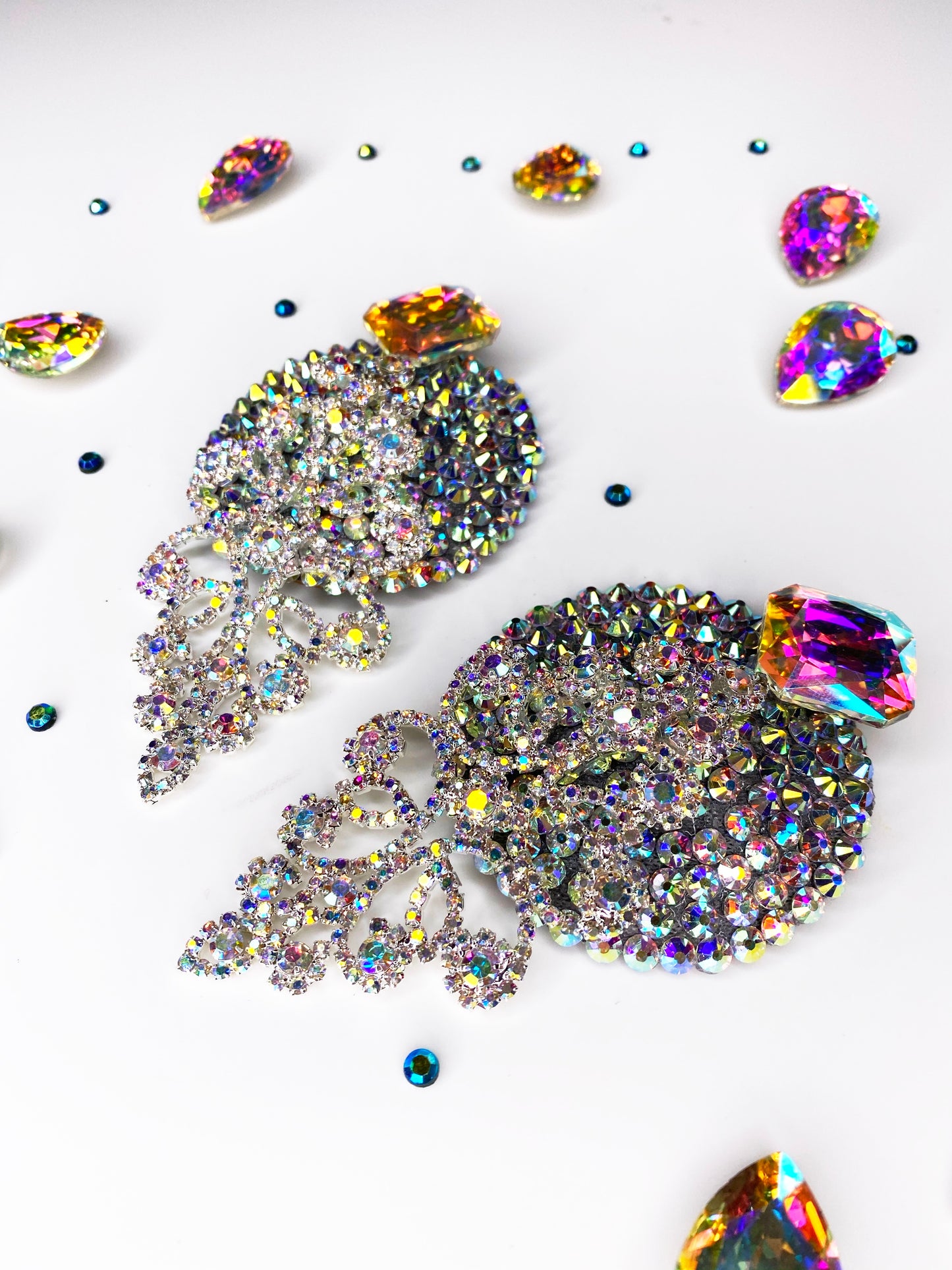 The Leading Lady earrings by Viva La Bombshell Burlesque feature iridescent rhinestones and intricate designs, displayed on white and surrounded by vibrant gemstones and crystals, adding a dazzling touch to your look.