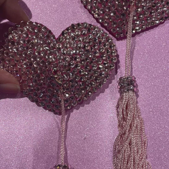 A hand holding pink rhinestoned pasties with pink tassels on a glittery pink background. 