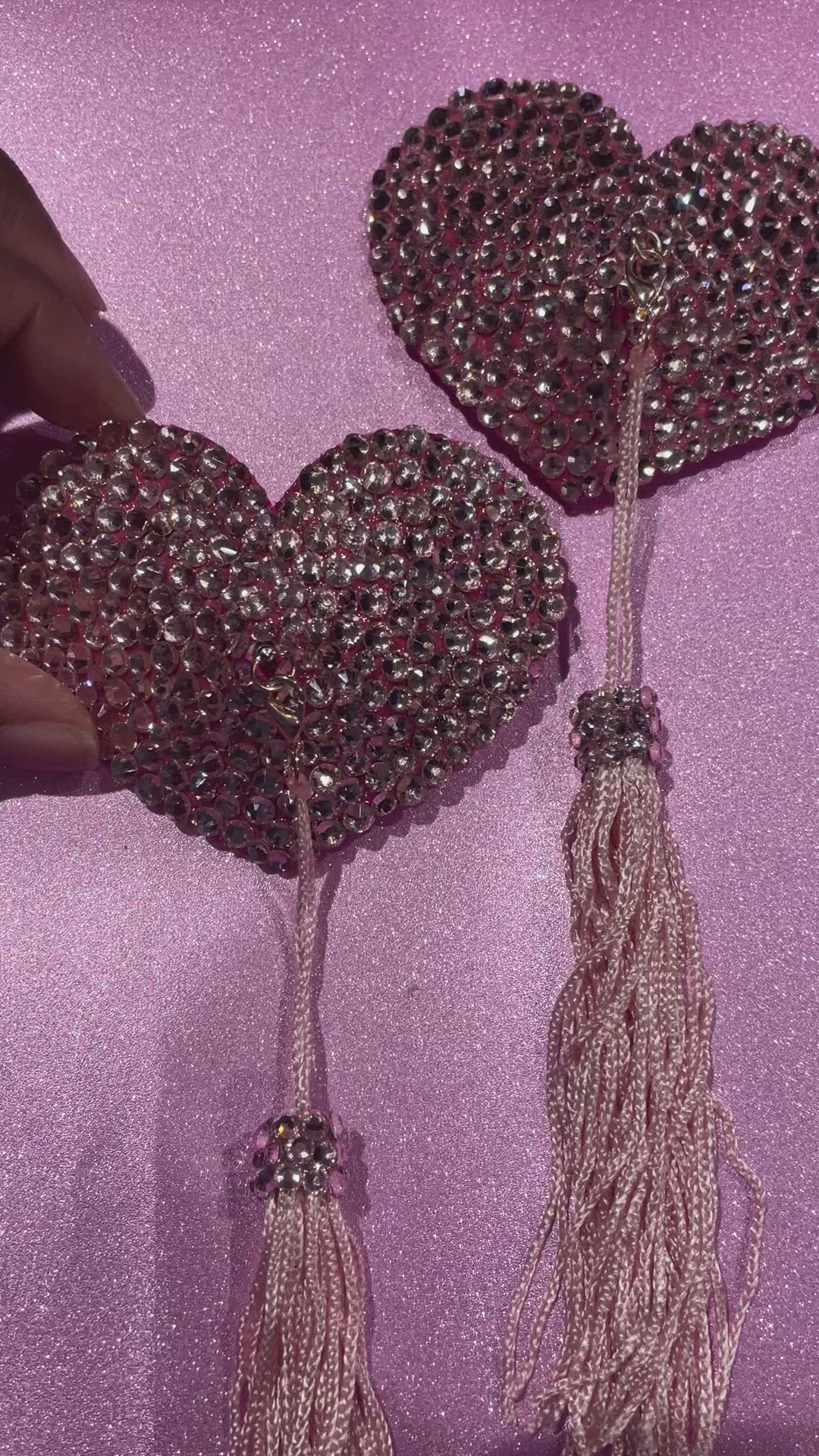 A hand holding pink rhinestoned pasties with pink tassels on a glittery pink background. 