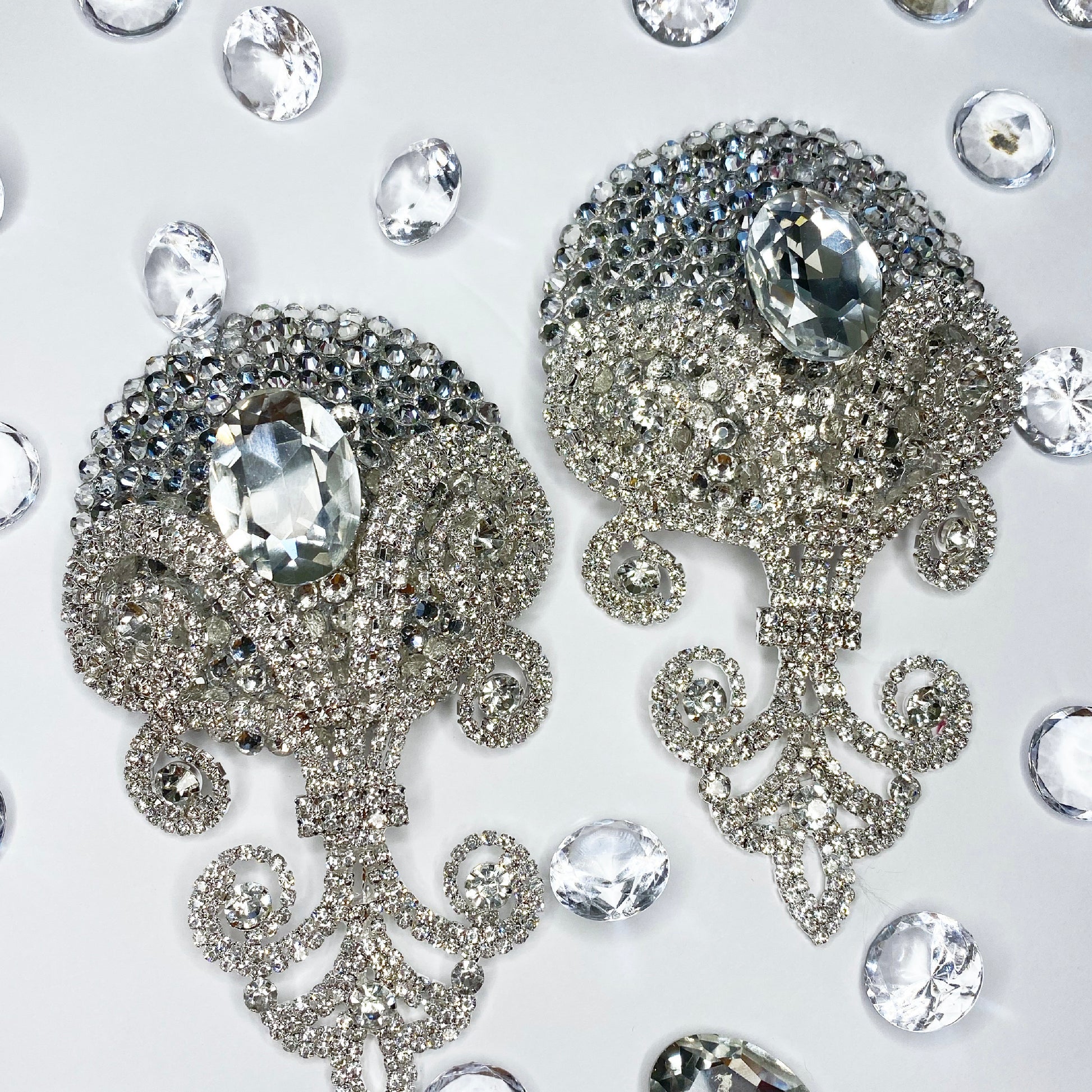 Two ornate silver Viva La Bombshell Burlesque Showstopper Pasties brooches with large oval crystals are displayed on a white background, surrounded by scattered clear gemstones.