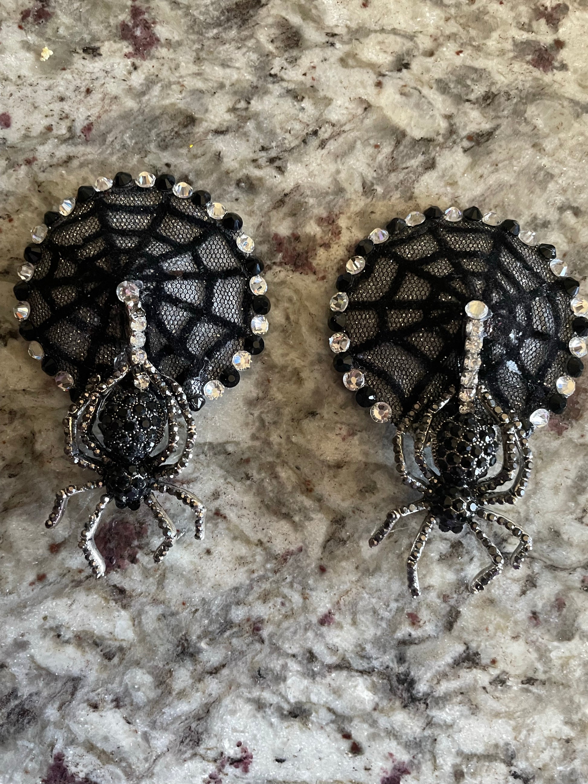 Viva La Bombshell Burlesque's Mistress of the Dark Pasties feature black spider figures on sparkling rhinestone webs, evoking Elvira style—perfect for a dramatic burlesque look. Shown on a light mottled surface.