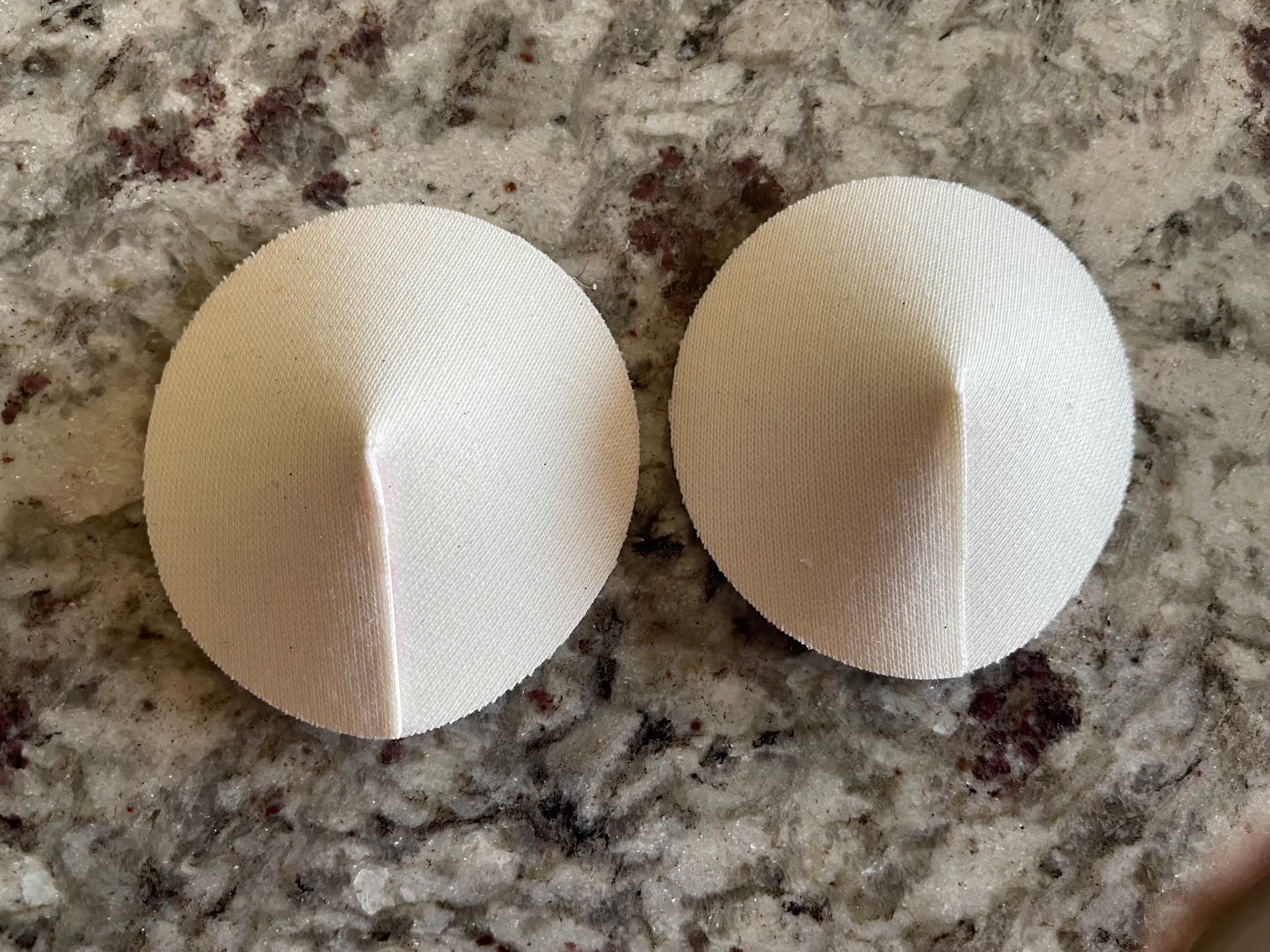 Two white DIY Pastie Base pads by Viva La Bombshell Burlesque, dome-shaped with a central seam, are placed side by side on a speckled gray countertop.