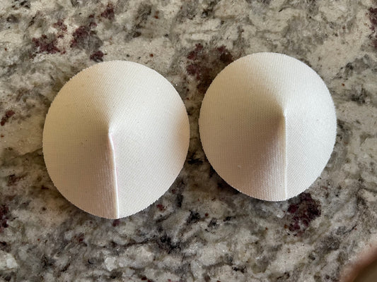Two white DIY Pastie Base pads by Viva La Bombshell Burlesque, dome-shaped with a central seam, are placed side by side on a speckled gray countertop.