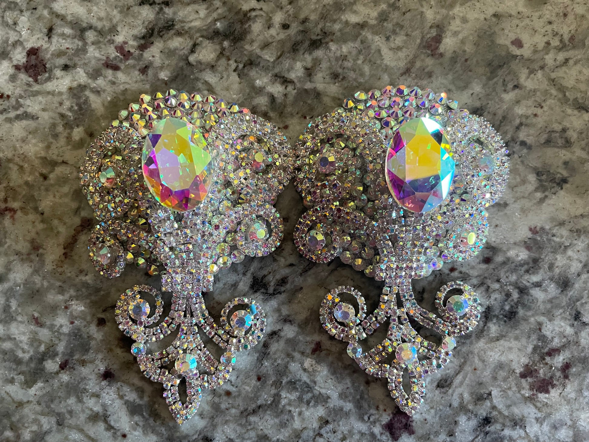 A pair of Showstopper Pasties by Viva La Bombshell Burlesque, featuring ornate iridescent rhinestones and large oval gems with intricate swirling designs, elegantly displayed on a marbled gray surface.