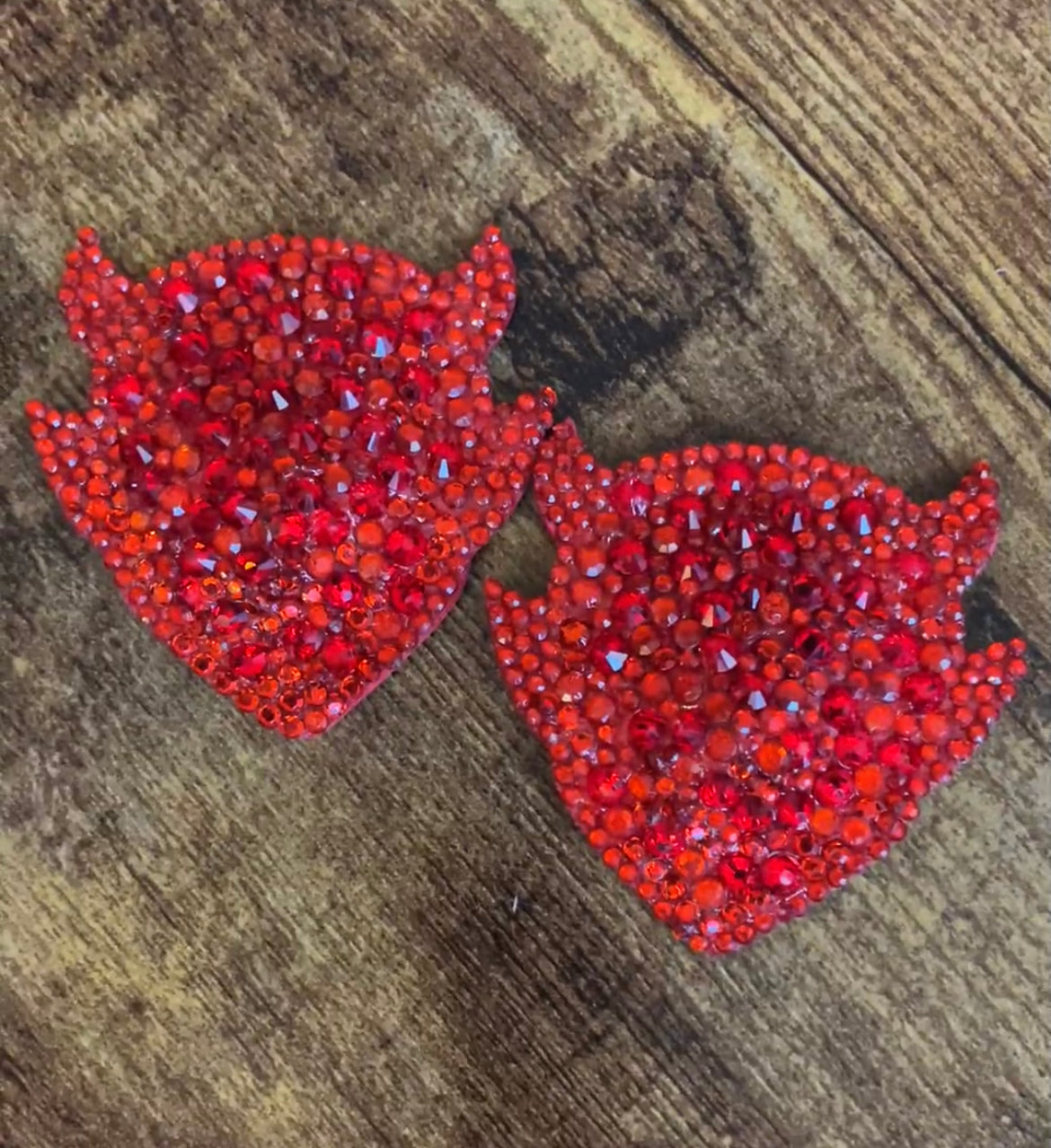 Two red H.I.M Pasties (Devil Silhouette) by Viva La Bombshell Burlesque, covered in rhinestones and shaped like horned devil faces, are displayed side by side on a wooden surface.