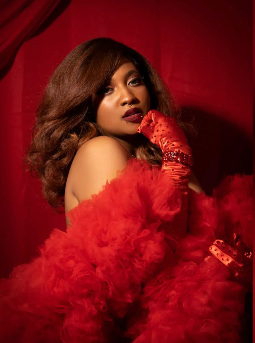 A woman with voluminous brown hair poses confidently against a red backdrop, wearing The Diva - Tulle Boa by Viva La Bombshell Burlesque and red embellished gloves, with bold makeup and an intense gaze.